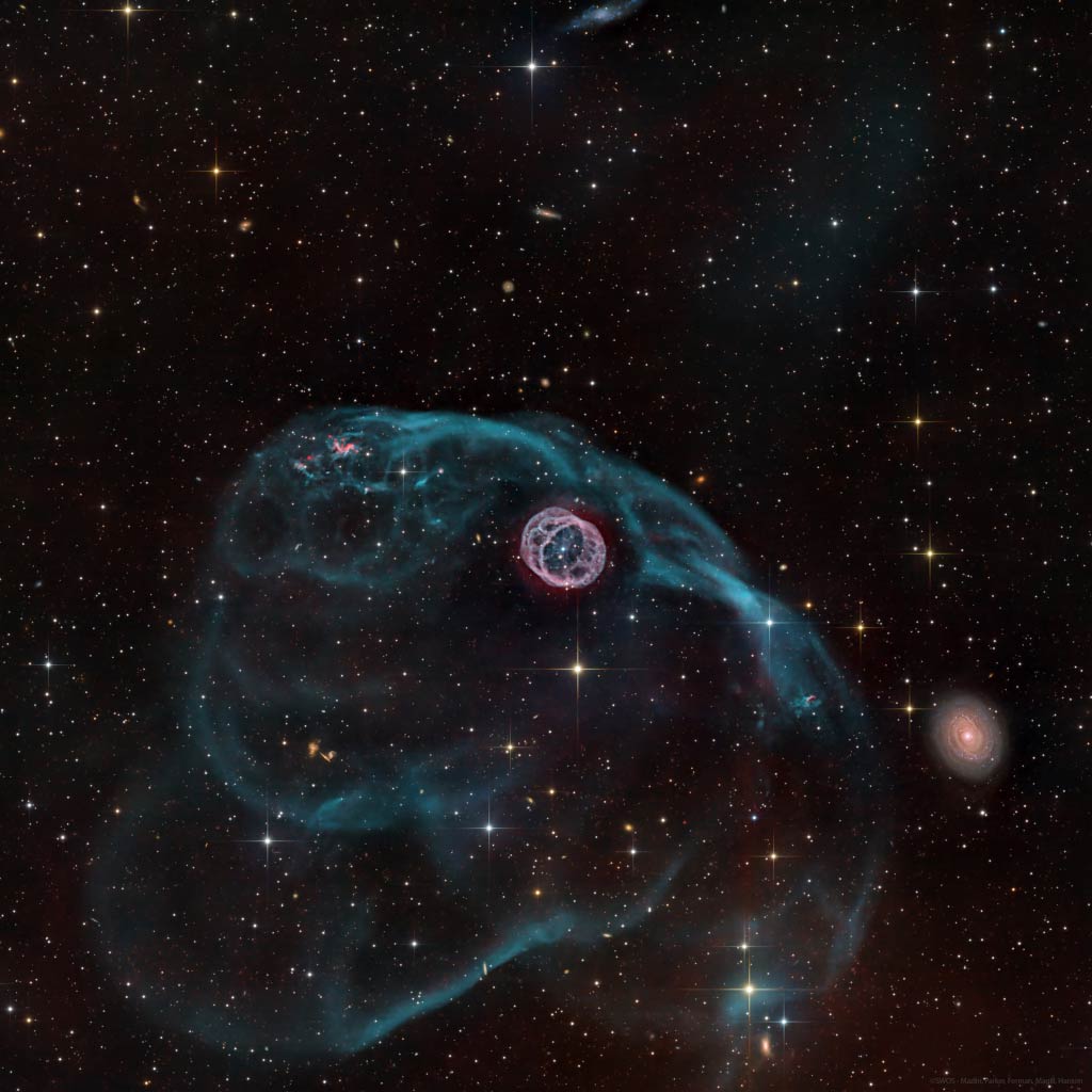 Mitten im Bild ist eine kompakte rosatote Struktur, die an zwei Kreise erinnert, die mit Speichen verbunden sind. Dahinter zieht eine türkisblaue Nebelstruktur. Rechts ist eine helle Spiralgalaxie.