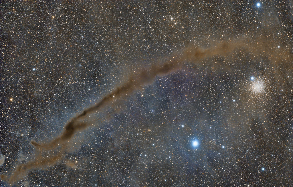 Ein dunkler Nebelstreif zieht vor einem Himmel voller Sterne von links unten diagonal nach oben. Rechts schimmert ein Kugelsternhaufen, der aber viel weiter entfernt ist als das dunkle Dingsbums.