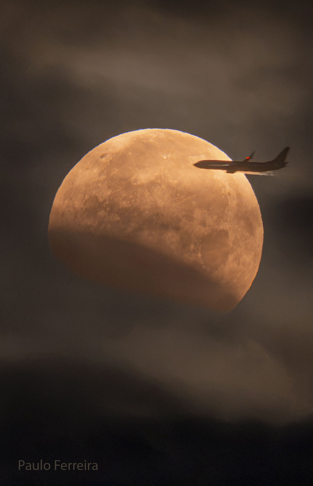 Der teilweise verfinsterte Mond füllt beinahe das Bild. Von rechts fliegt ein Flugzeug davor vorbei. Der dunkle Mondschatten ist links unten.
