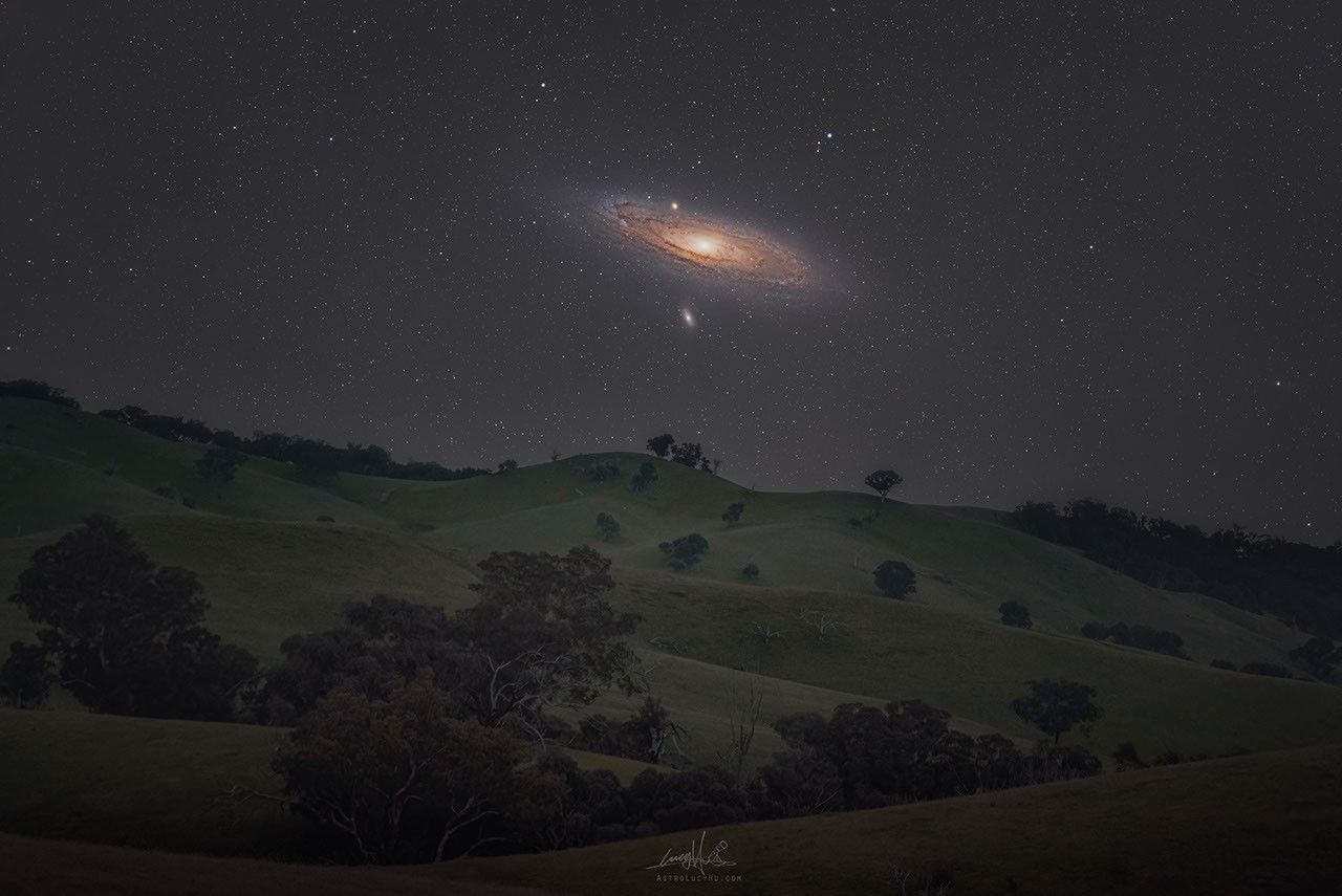 Die nächtliche Landschaft mit Hügeln und Bäumen liegt in Australien. Über dem Horizont steht die Andromeda-Galaxie M31. Auch ihre Begleiterinnen M32 und M110 sind gut erkennbar. Die Galaxie erscheint im Vergleich zur Landschaft riesig. Sie ist so breit wie sechs Vollmonde.