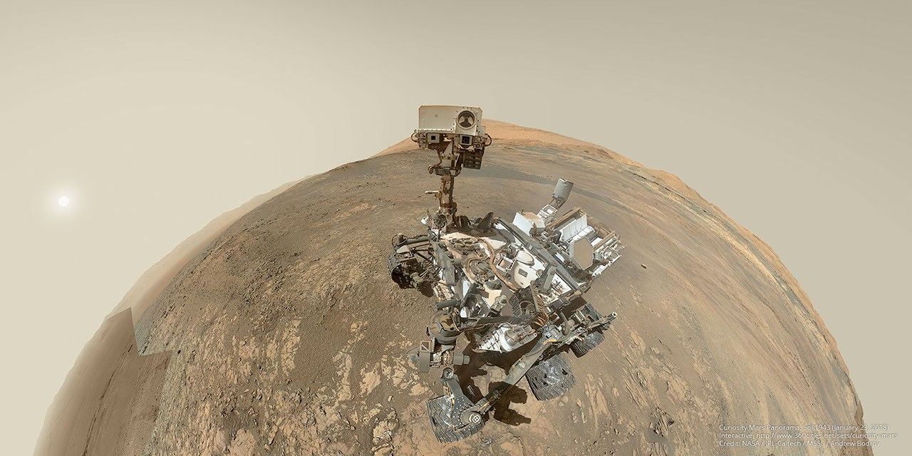 Selbstbildnis eine fahrenden Roboters auf der Marsoberfläche in Fischaugenoptik. Die Planetenoberfläche ist mit braunem Sand und hellen Steinen bedeckt und ist in einen Halbkreis gekrümmt. Am hellbraunen Himmel darüber steht links die schwache Sonne.